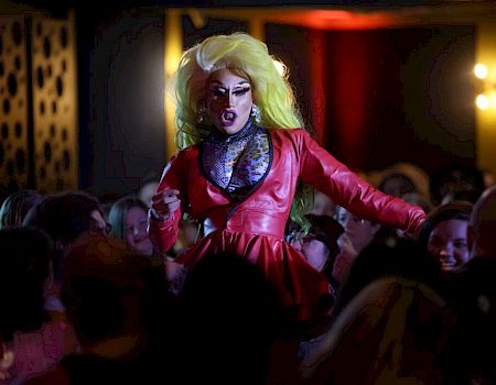 A performer with vibrant yellow hair and a red outfit entertains a crowd in what appears to be a lively indoor venue.