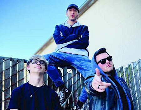 Three individuals pose outdoors, with one sitting on a fence and the other two standing in front.