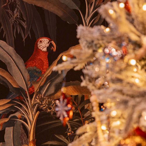 A parrot is perched among tropical foliage, with a decorated and lit Christmas tree in the foreground, creating a festive atmosphere.