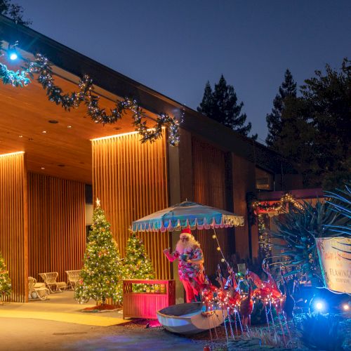 A Christmas-themed outdoor scene with decorated trees, garlands, and lighting, featuring Santa under an umbrella in a festive setup.