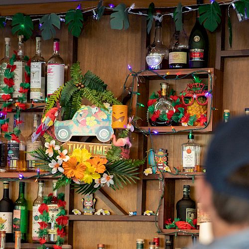 A decorated tropical-themed bar with various liquor bottles, colorful lights, flowers, and a 