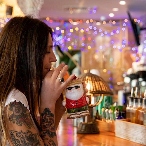 A woman with tattoos holds a Santa Claus figurine in a decorated bar or cafe with colorful lights in the background.