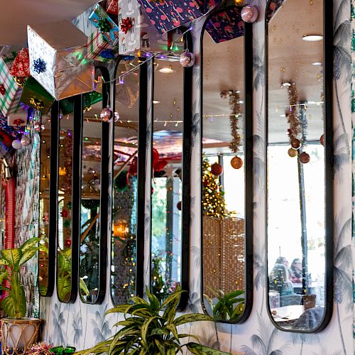 The image shows a decorated interior with several tall, narrow mirrors on the wall, plants, festive decor, and ornaments hanging from the ceiling.