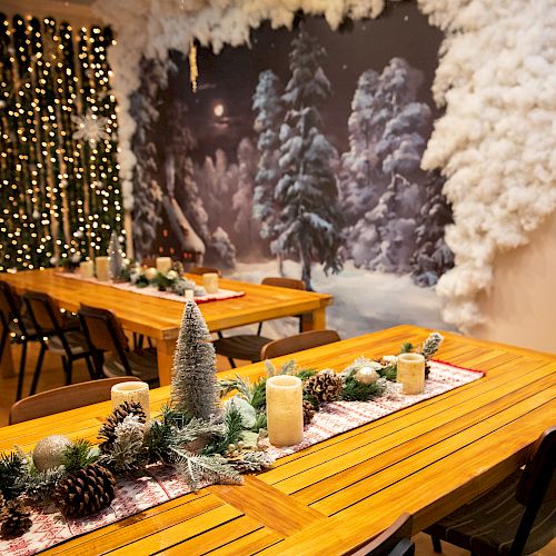 A festively decorated room with wooden tables, candles, pinecones, and greenery, set against a snowy forest backdrop and string lights.