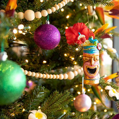 A decorated Christmas tree with colorful ornaments, including a purple ball, a green ball, and a detailed tiki mask ornament, lit by small white lights.