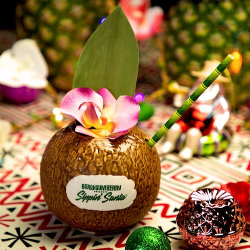 A tropical-themed drink in a coconut shell with a straw and flower garnish on a colorful patterned table, surrounded by fruit and festive decorations.