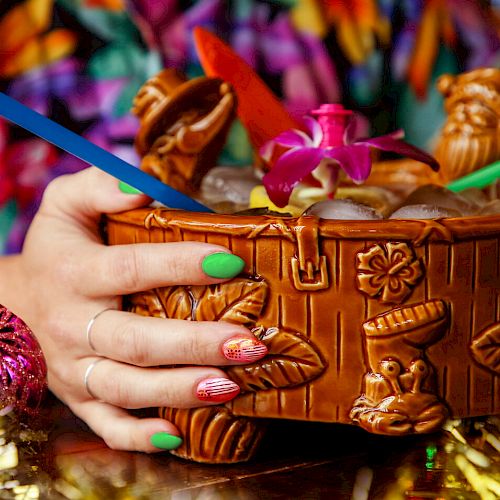 A person with colorful nails holds an ornate tiki bowl containing a tropical drink, with straws and a purple flower as decoration, surrounded by festive decor.