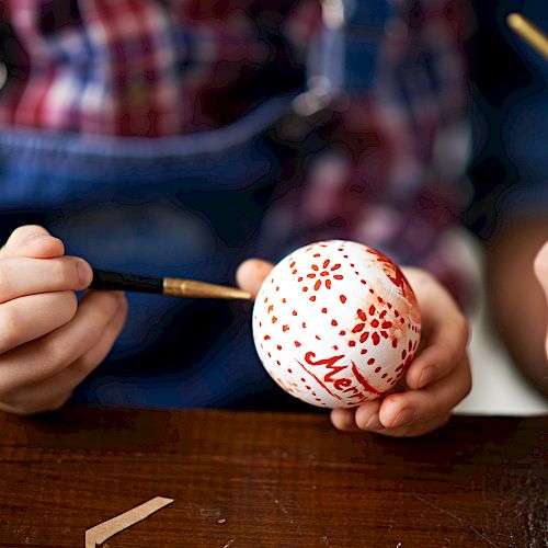 A person is hand-painting a Christmas ornament with red paint, holding it carefully with one hand while using a brush in the other hand.