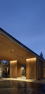 A modern, well-lit building entrance with a large overhang and glass doors, set against a dim evening sky.