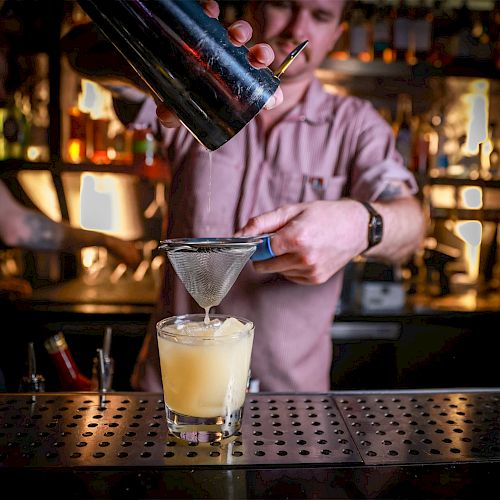 A bartender is carefully straining a cocktail into a glass filled with ice, standing behind a bar with various bottles on the shelves.