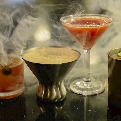 The image shows a group of four cocktails on a bar, surrounded by a misty, smoky effect, with a cherry garnish and lime wedge visible.