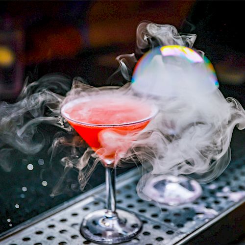 A red cocktail in a martini glass is emitting smoke or vapour, with a bubble decoration on top, placed on a bar counter.