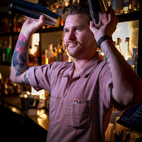 A bartender with tattooed arms is smiling and holding two shaker tins above his head, standing behind a bar with various bottles in the background.