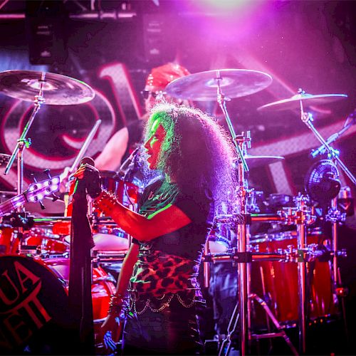 A band is performing on stage with vibrant lighting; the drummer is visible behind the drum set.