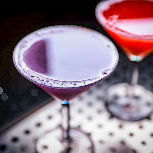 The image shows two cocktail glasses filled with purple and red drinks, placed on a metal surface dotted with small holes.