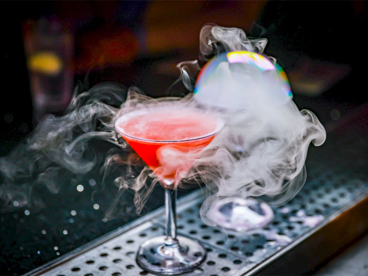 A vibrant red cocktail in a glass sits on a bar counter, surrounded by dramatic swirling smoke effects, creating an intriguing and stylish presentation.