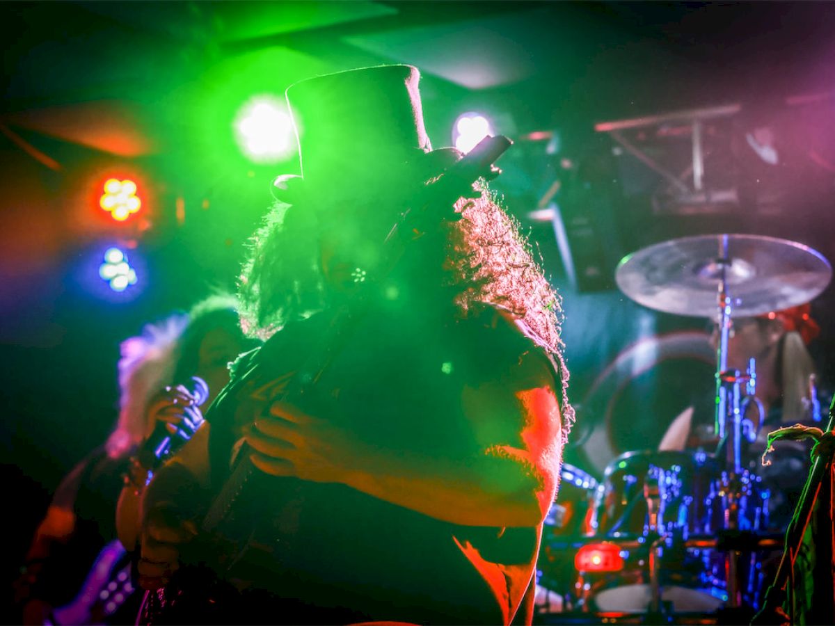 A musician with long hair and a top hat plays guitar under colorful stage lights, with a drum set visible in the background.