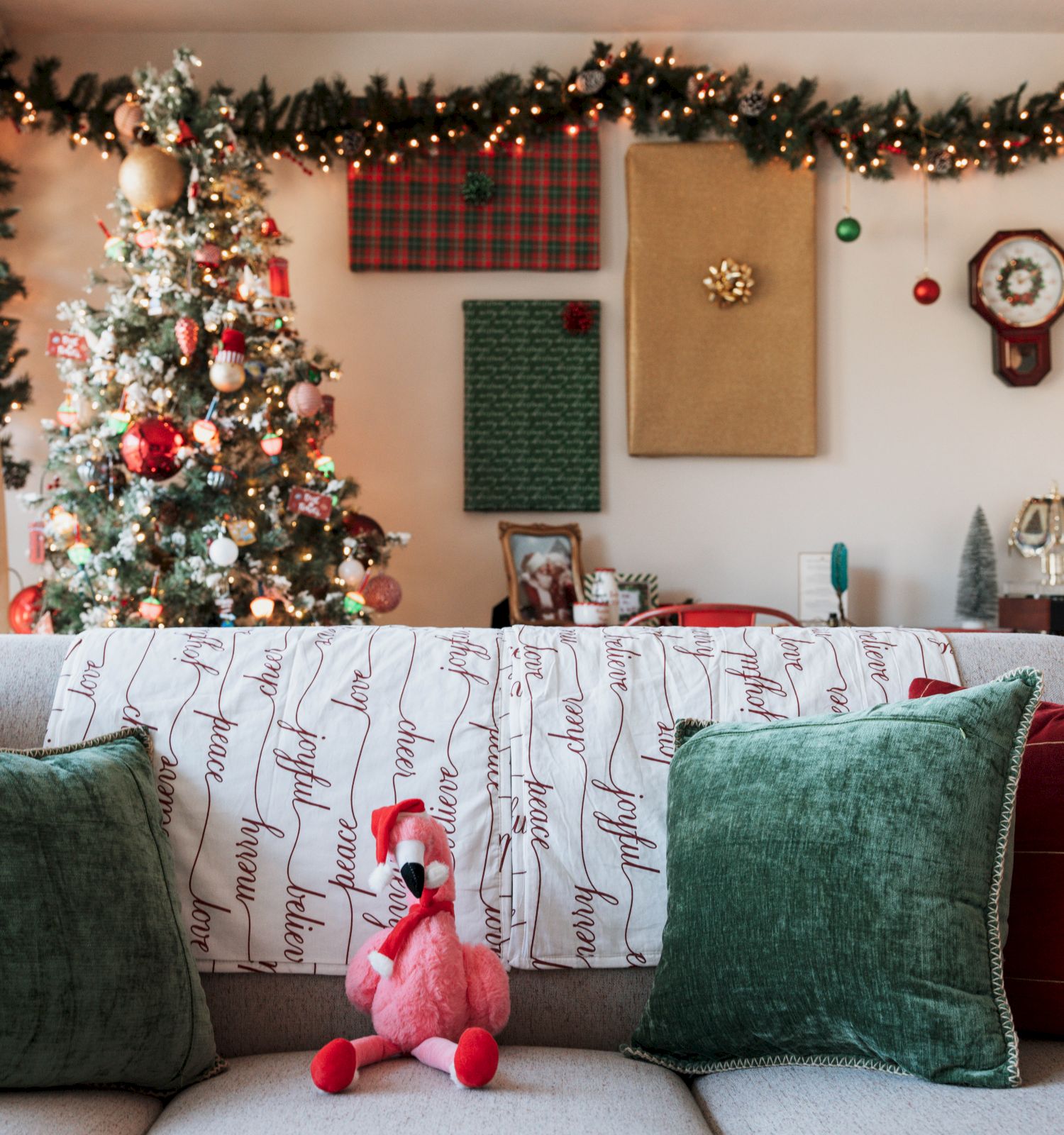 A living room decorated for Christmas with a tree, garlands, ornaments, and festive lights. A sofa with green and red pillows faces the tree.