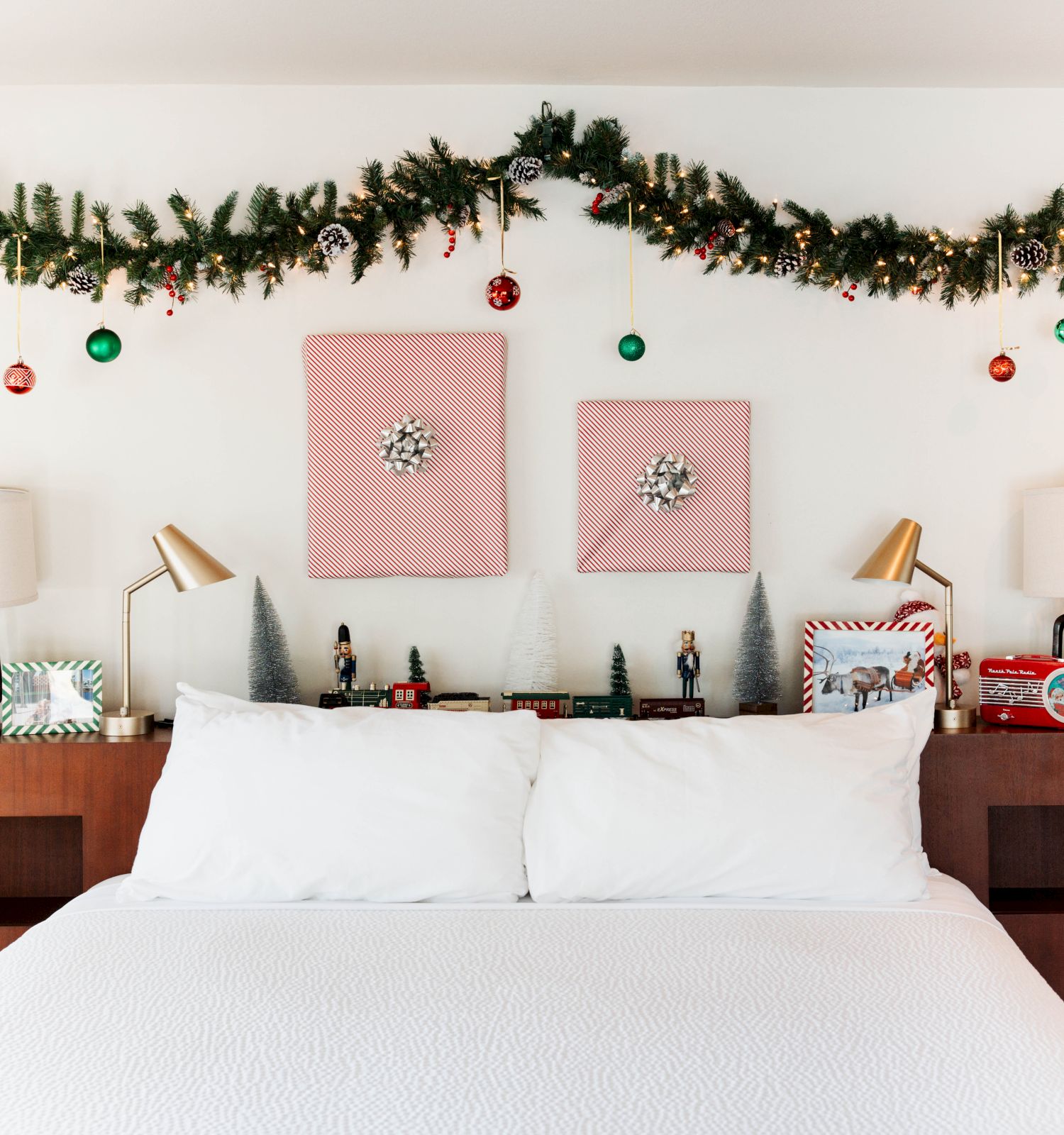 A festive bedroom with a garland, red bows, ornaments, and a decorated headboard above a bed with white pillows and lamps on each side.