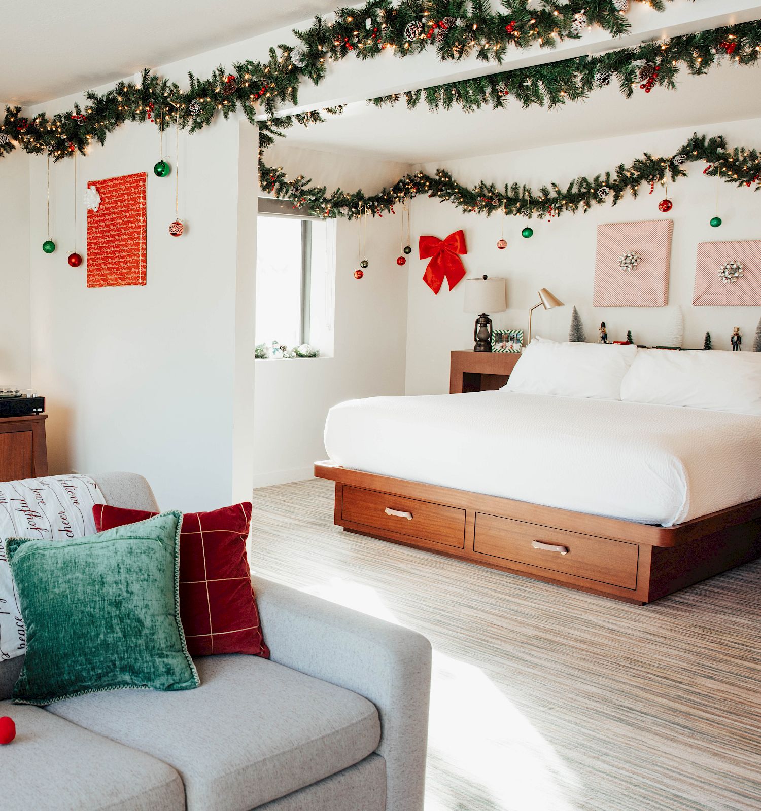 A festive room decorated for Christmas with garlands, lights, a tree, and red and green accents, featuring a bed, sofa, and small desk area.