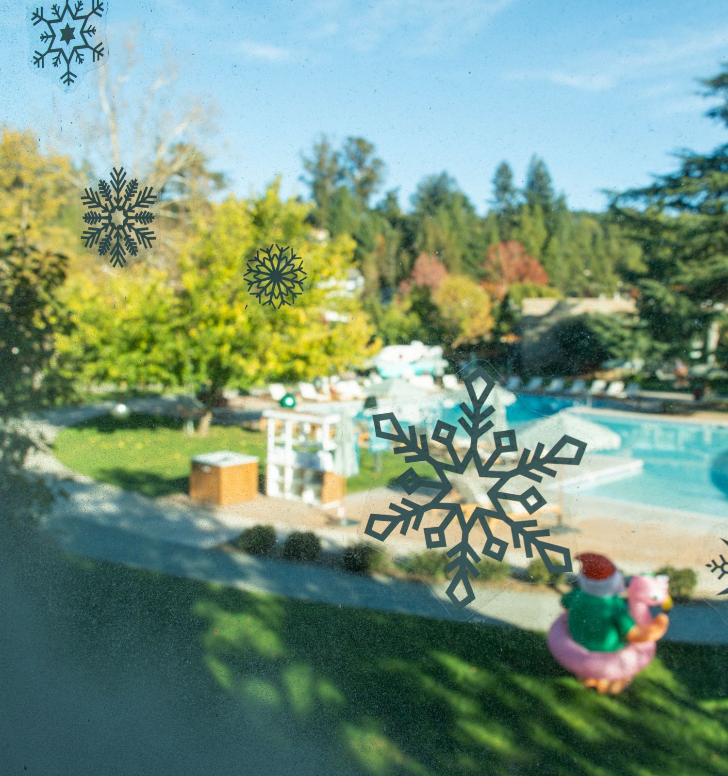 A poolside outdoor area with holiday decorations seen through a window adorned with snowflake stickers, surrounded by trees and greenery.