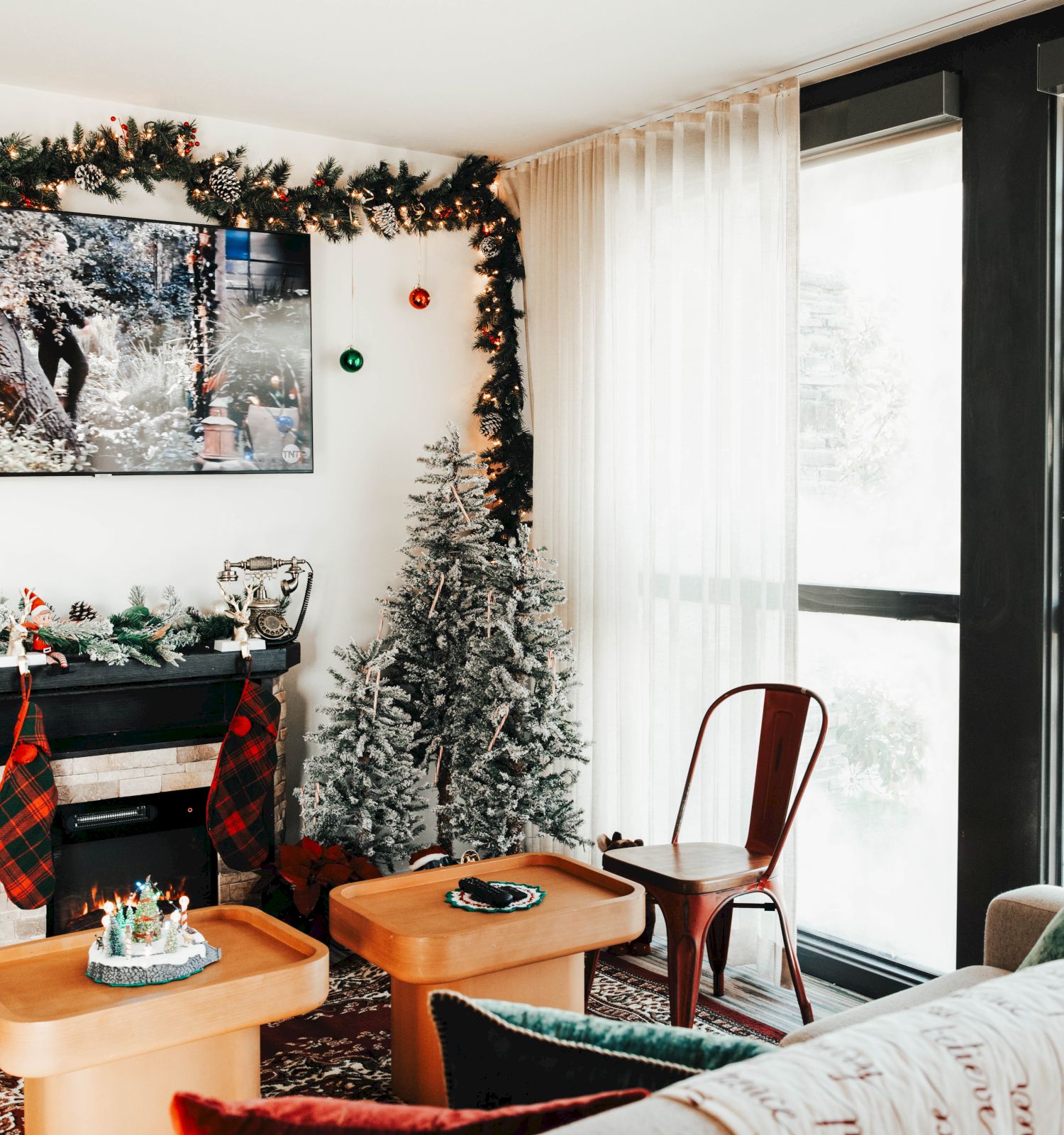 A cozy living room with festive decorations, including a Christmas tree, garland, and a merry fireplace; a TV shows a holiday scene.