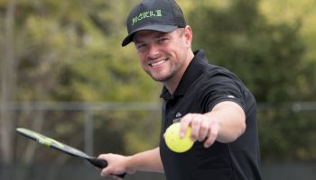 A person is on a tennis court, holding a tennis racket and a ball while smiling at the camera, wearing a black cap and shirt.