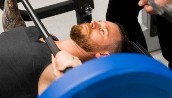 A man is lying on a bench, lifting a barbell with blue weight plates, while another person stands nearby, possibly offering support or spotting him.