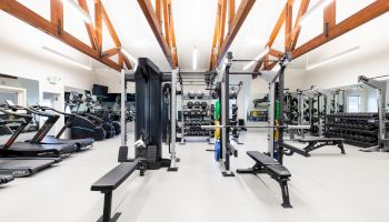 A modern gym with treadmills, weight benches, squat racks, and various exercise equipment, with wooden ceiling beams and bright lighting.