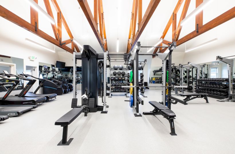 A modern gym with treadmills, weight benches, squat racks, and various exercise equipment, with wooden ceiling beams and bright lighting.