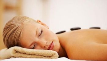 A person is lying down and appears to be receiving a hot stone massage on their back, with their eyes closed and head resting on a towel.