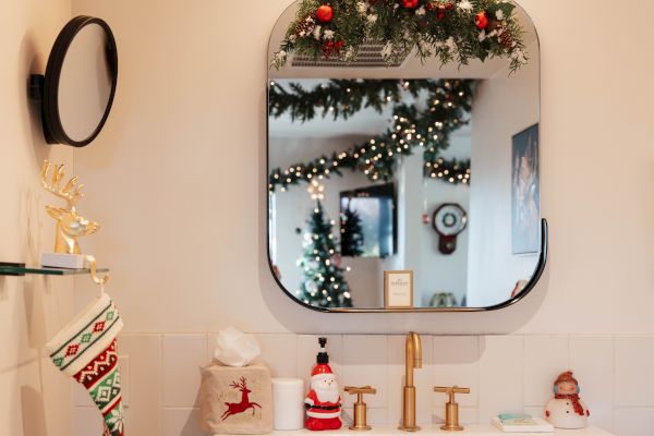 A bathroom mirror reflects a decorated Christmas tree with garlands. A stocking, snowman, and festive decor are on the counter and shelves.