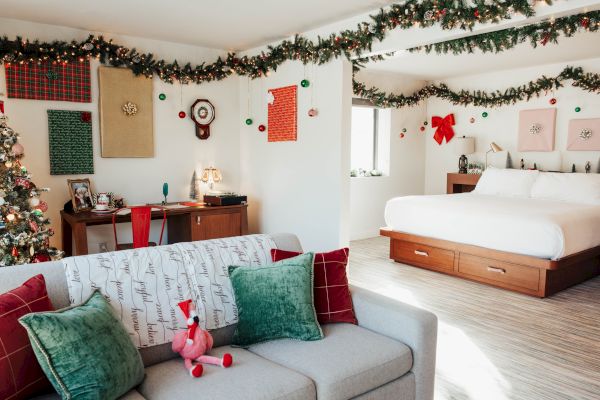 A festively decorated room with holiday garlands, a Christmas tree, a cozy bed, a couch with pillows, and a desk with seasonal decor.