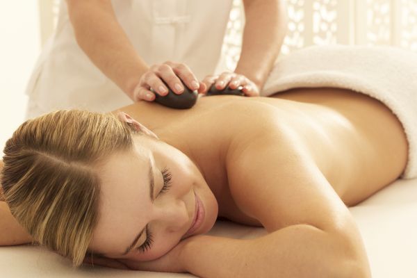 A person is lying on a massage table, receiving a hot stone massage on the back from a therapist, and appears to be relaxed.