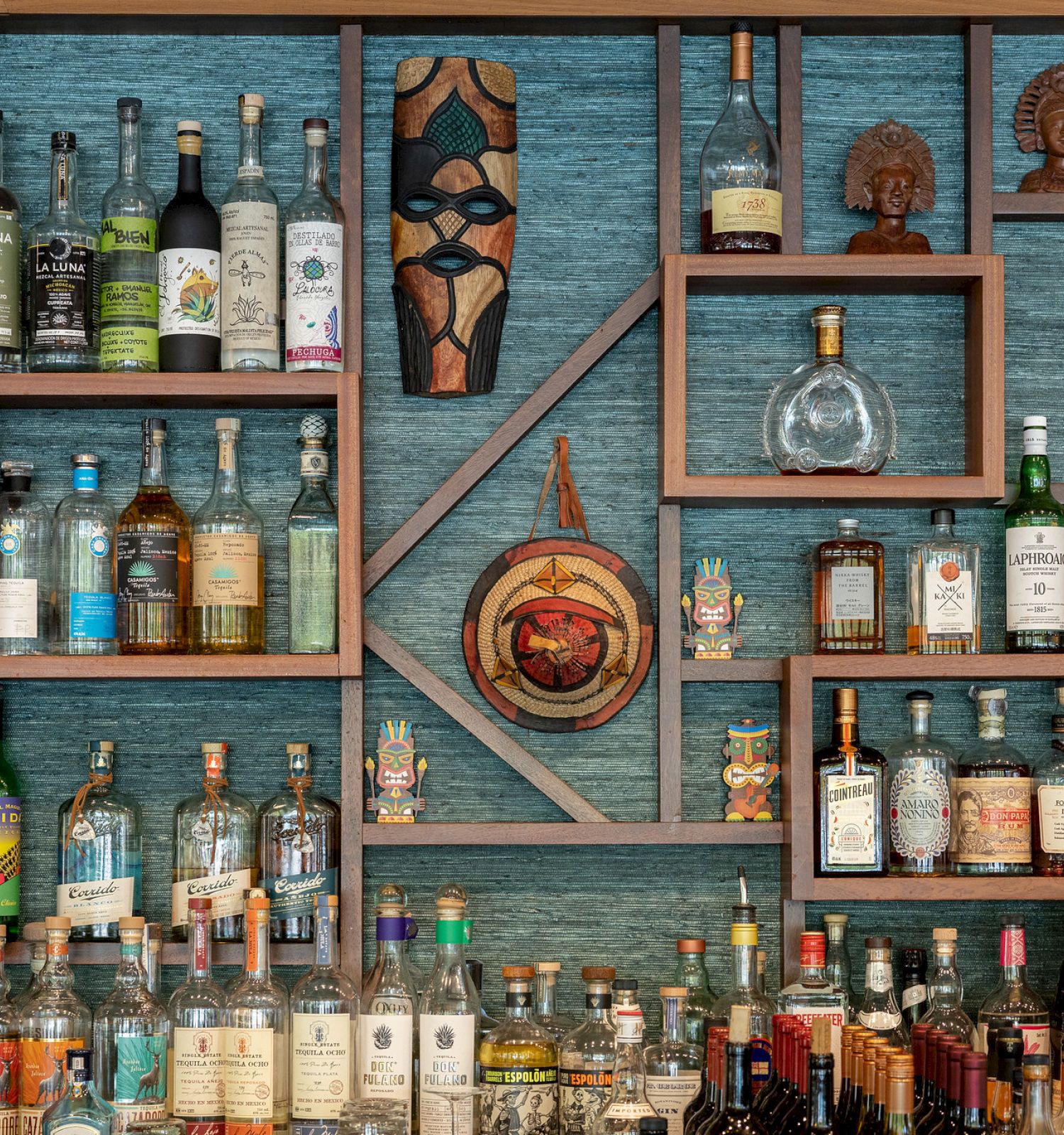 The image shows various bottles of alcohol on shelves arranged in a geometric pattern, bookended by traditional masks and statuettes.