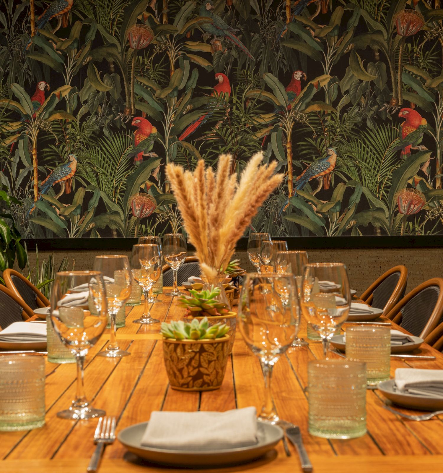 A dining table is elegantly set with glasses, plates, and napkins. The background features a tropical-themed wallpaper.