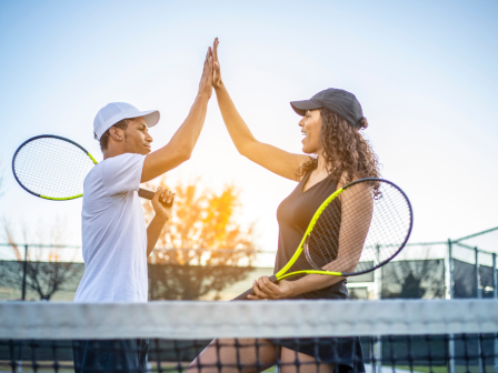 Two tennis players on a court celebrate a win with a high-five, both holding their racquets and smiling at each other.