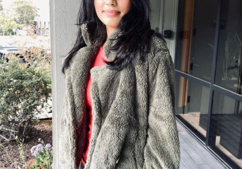 A woman stands outdoors wearing a fuzzy green jacket and red top, smiling softly with a building and greenery in the background.