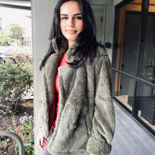 A woman stands outdoors wearing a fuzzy green jacket and red top, smiling softly with a building and greenery in the background.