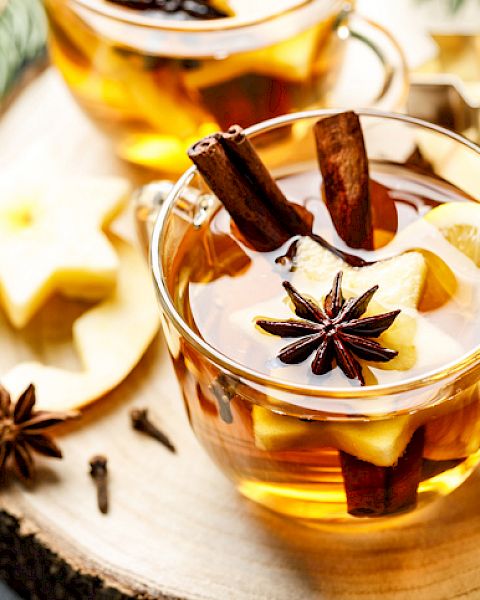 A warm beverage in a glass cup with cinnamon, star anise, and apple slices on a wooden surface.