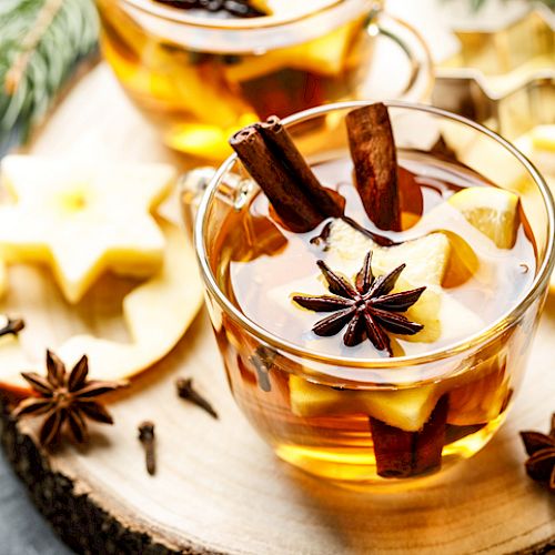 A warm beverage in a glass cup with cinnamon, star anise, and apple slices on a wooden surface.