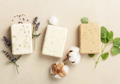 Three bars of soap are placed alongside sprigs of lavender, cotton, and green leaves on a light beige background.