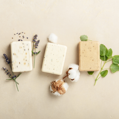 Three bars of soap are placed alongside sprigs of lavender, cotton, and green leaves on a light beige background.