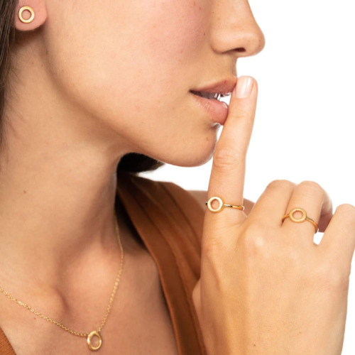 A person with gold jewelry (earring, ring, necklace) in a circular design, holding a finger to their lips in a shushing gesture.