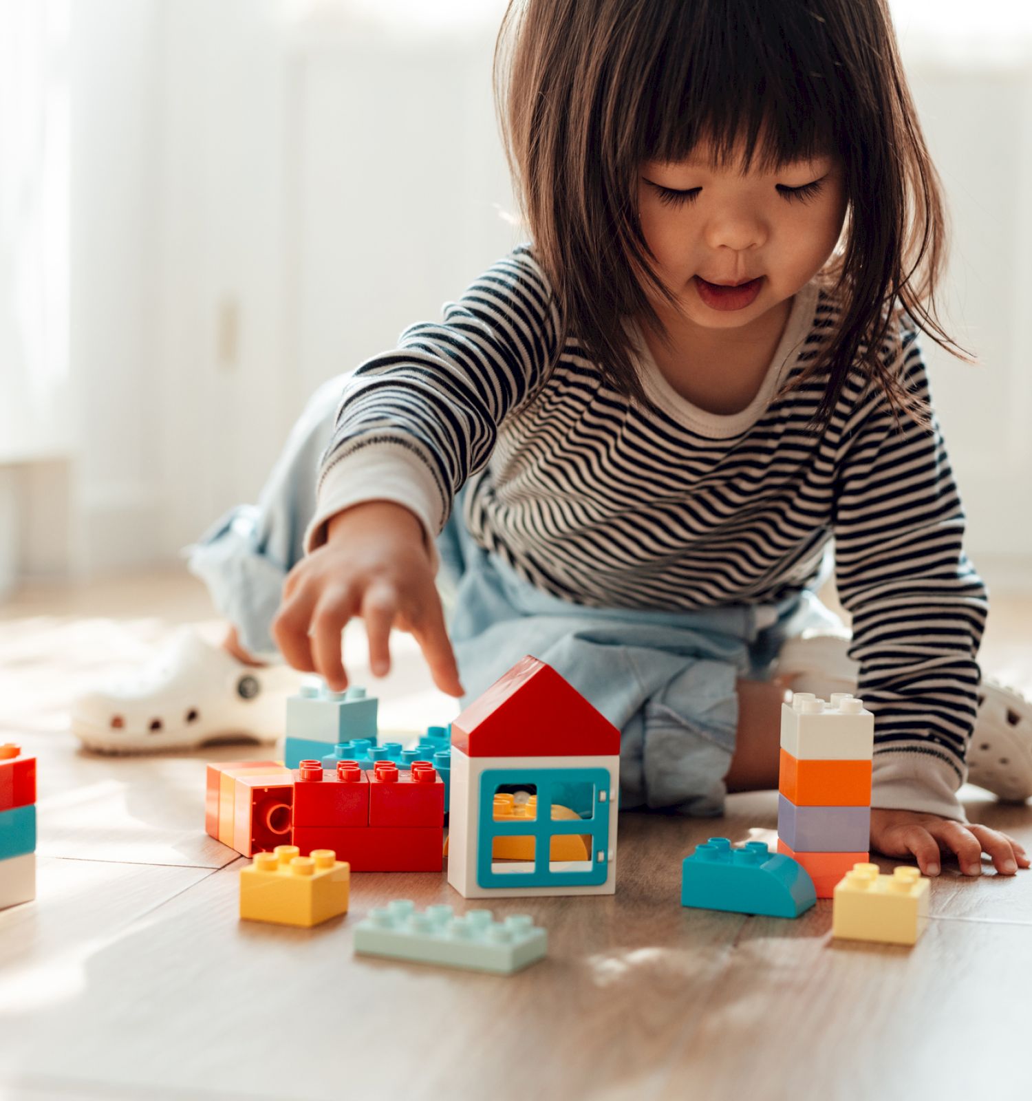 A child playing with colorful building blocks on a wooden floor, wearing a striped shirt and surrounded by natural light from a window.