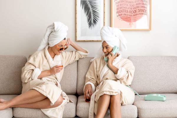 Two people in bathrobes with towels on their heads sit on a couch, one on the phone, both smiling, with drinks nearby, enjoying a relaxing time.