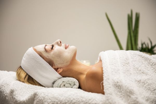 A person is lying down, covered in a white towel, with a face mask on and a headband, suggesting a spa or relaxation setting.