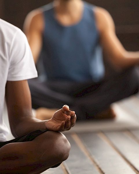 Several people are seated on yoga mats, meditating with one individual in a white shirt and the others out of focus.