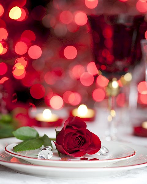 A romantic dinner setting with a red rose on a plate, surrounded by red lights, candles, and glasses in the background.