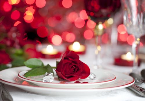 A romantic dinner setting with a red rose on a plate, surrounded by red lights, candles, and glasses in the background.